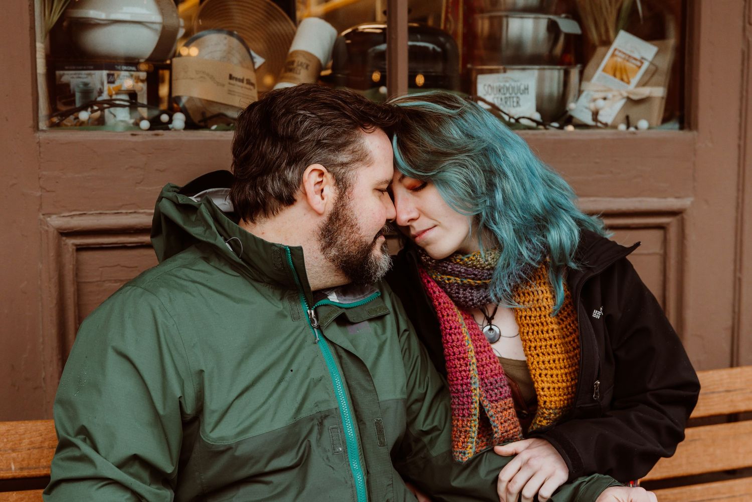 Couple with blue-haired woman and bearded man touching foreheads tenderly on an outdoor bench.