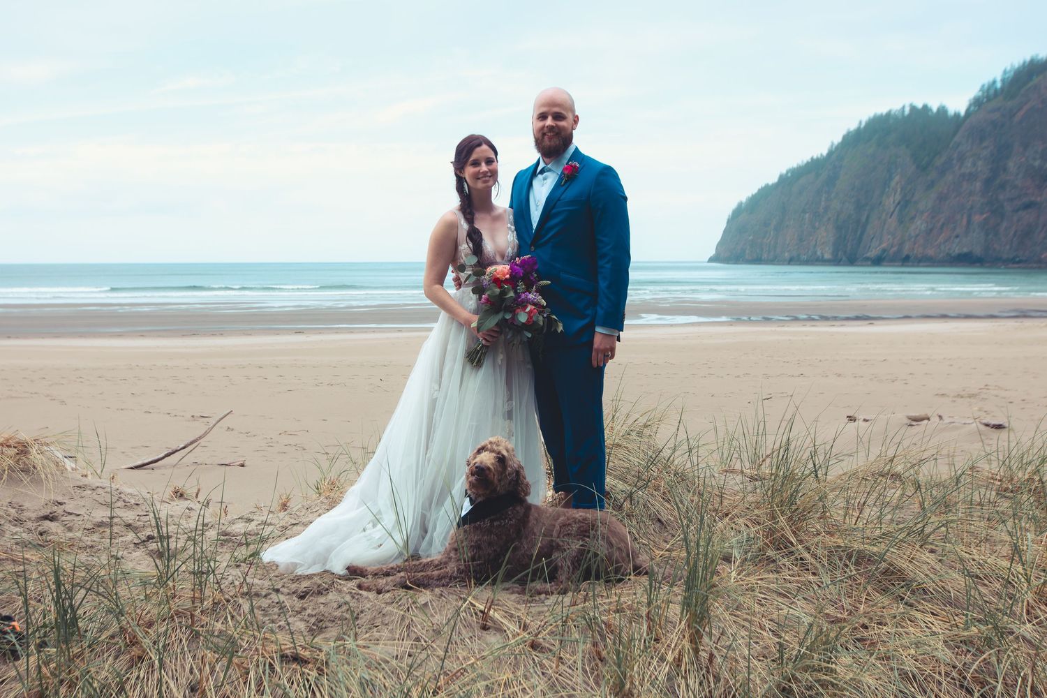 Newlyweds and their fluffy brown dog sitting in beach grass on their wedding day.