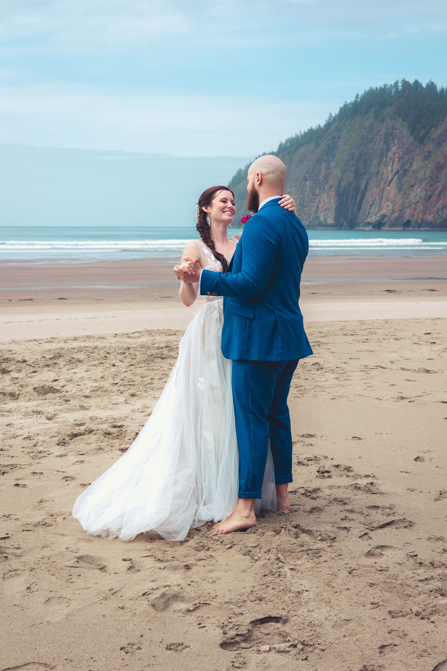 Newlyweds share a joyful moment barefoot on beach, groom in blue suit holding small item, cliffs behind.