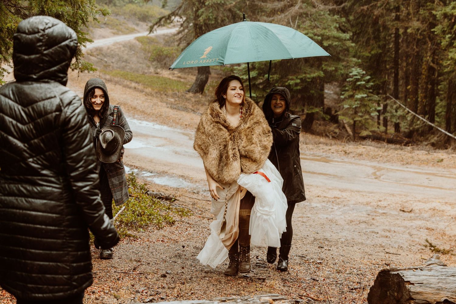 Bride in fur wrap laughs while guests help carry her dress on rainy forest path.