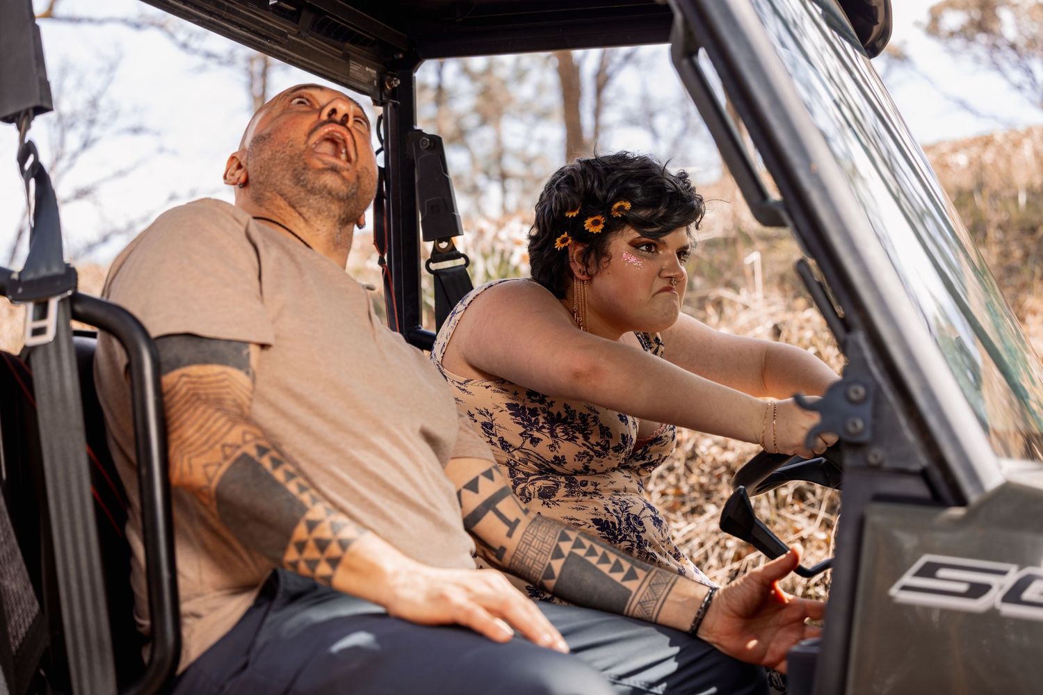 Man and woman sitting in off-road vehicle, woman in floral dress pointing forward.
