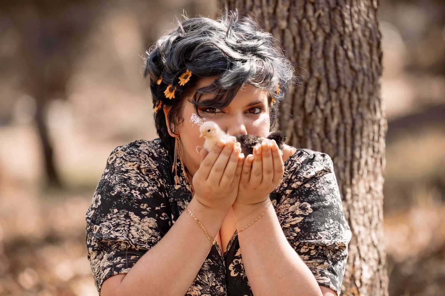 Woman with dark curly hair cups a white dove in her hands close to her face while leaning against a tree outdoors.