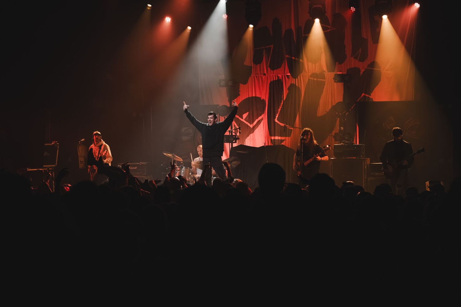 Rock band on stage with arms raised under dramatic red and orange stage lighting with crowd in foreground.