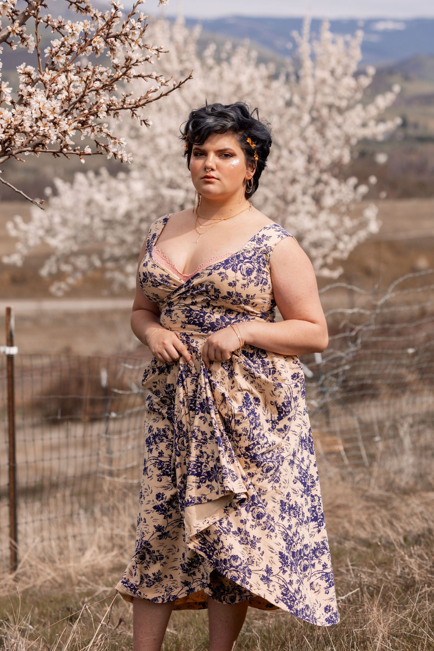 Woman in blue floral midi dress posing near blooming white tree in rural spring landscape.
