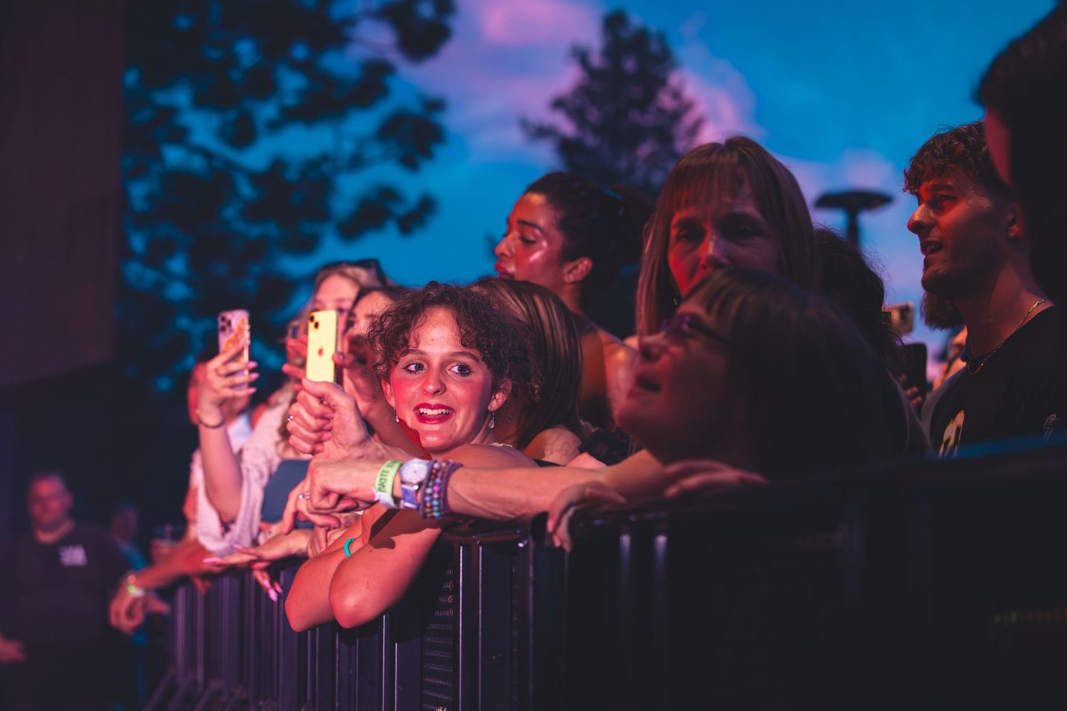 Excited concert fans lean on a barrier at an outdoor music event at dusk.