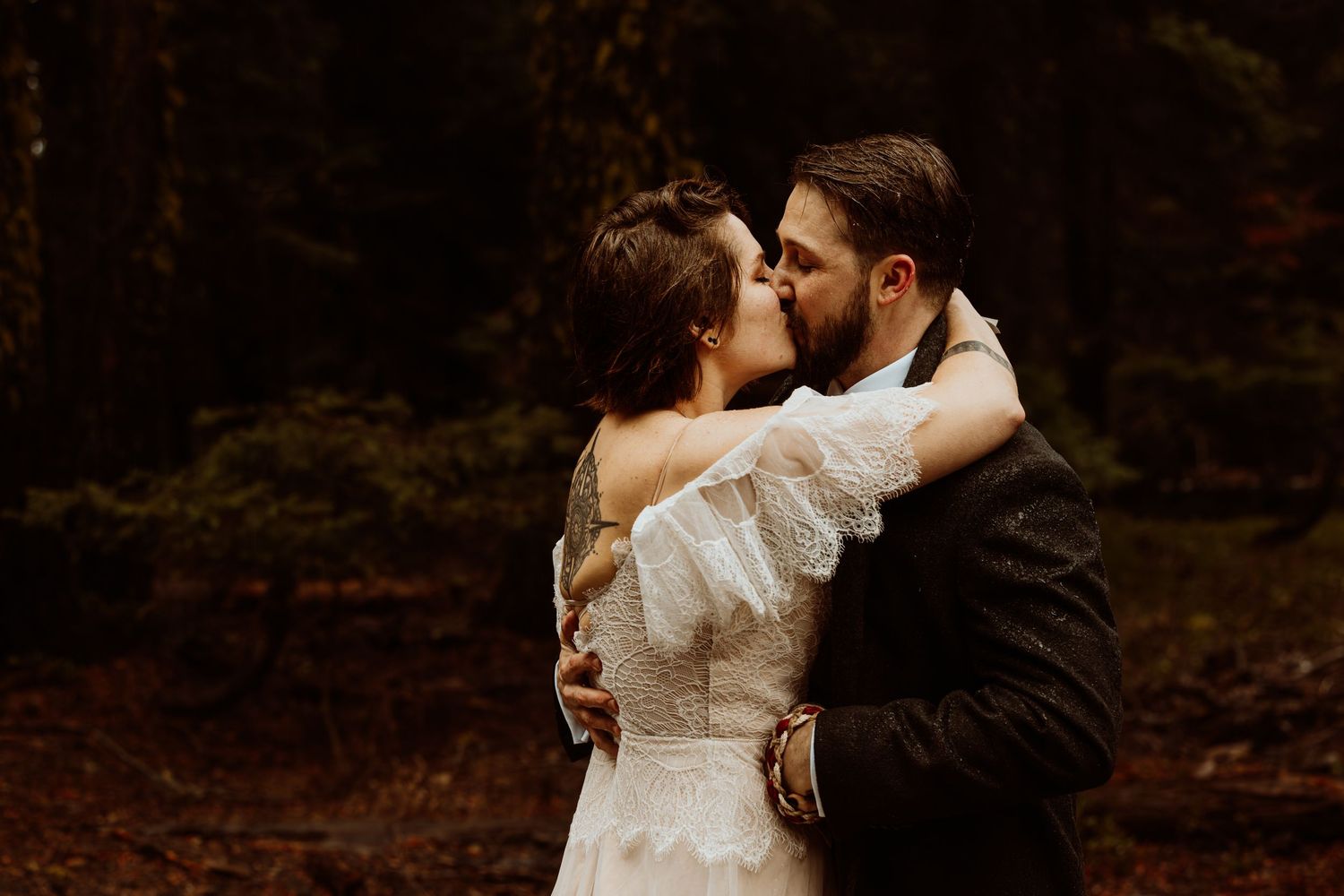 Bride and groom embracing with a tender kiss among tall dark trees in a forest wedding.