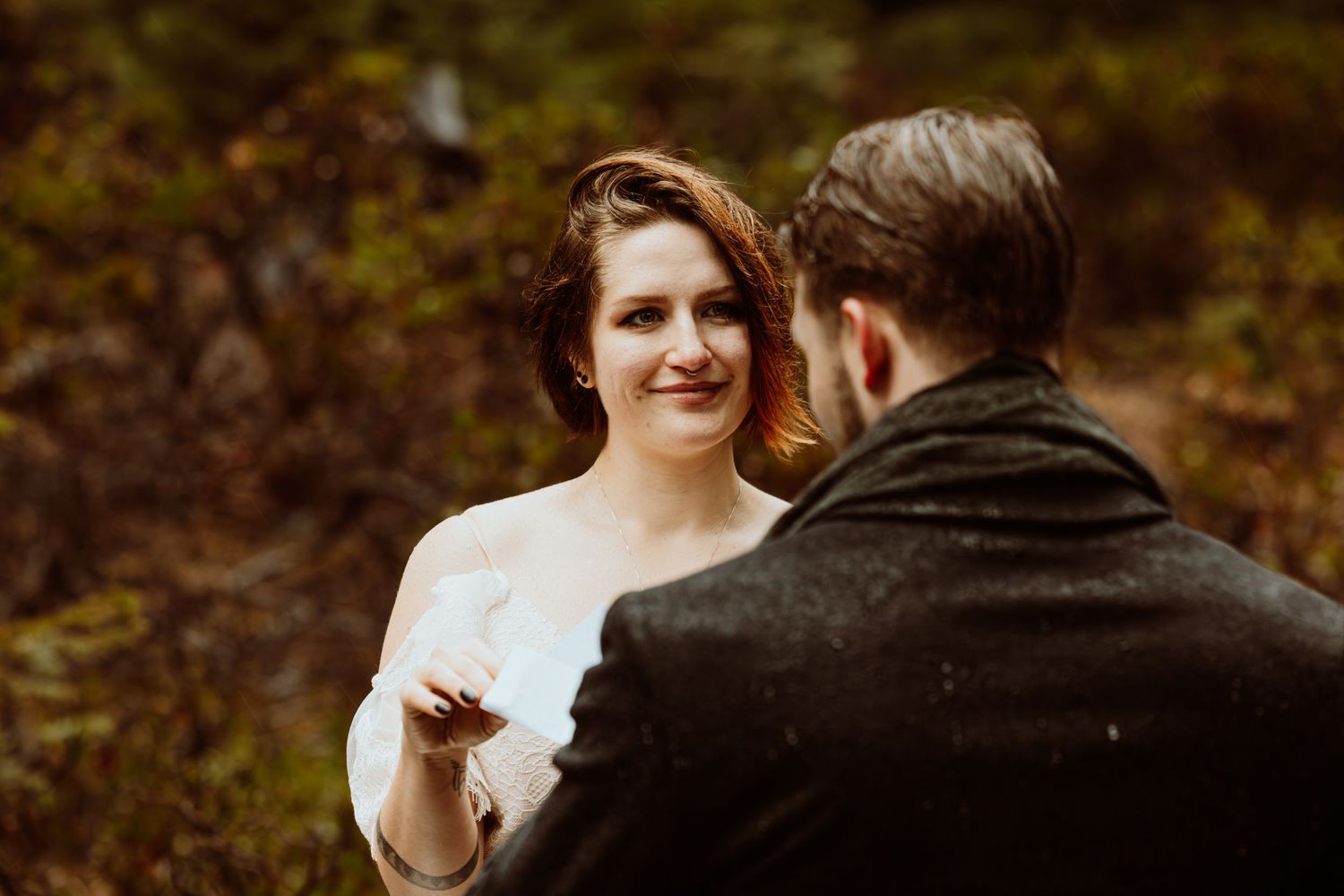 Bride smiles tenderly at groom during intimate rainy forest elopement ceremony in autumn setting.