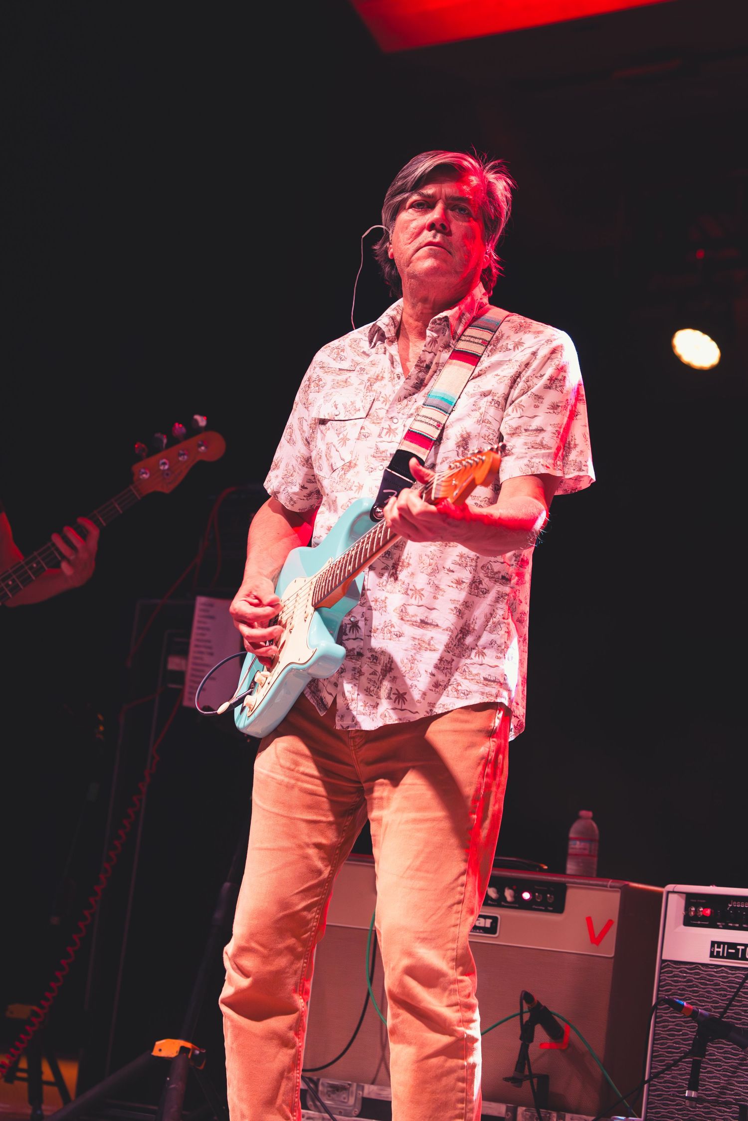 Guitarist performing live on stage playing a light blue Fender Stratocaster wearing a floral shirt under red stage lighting.
