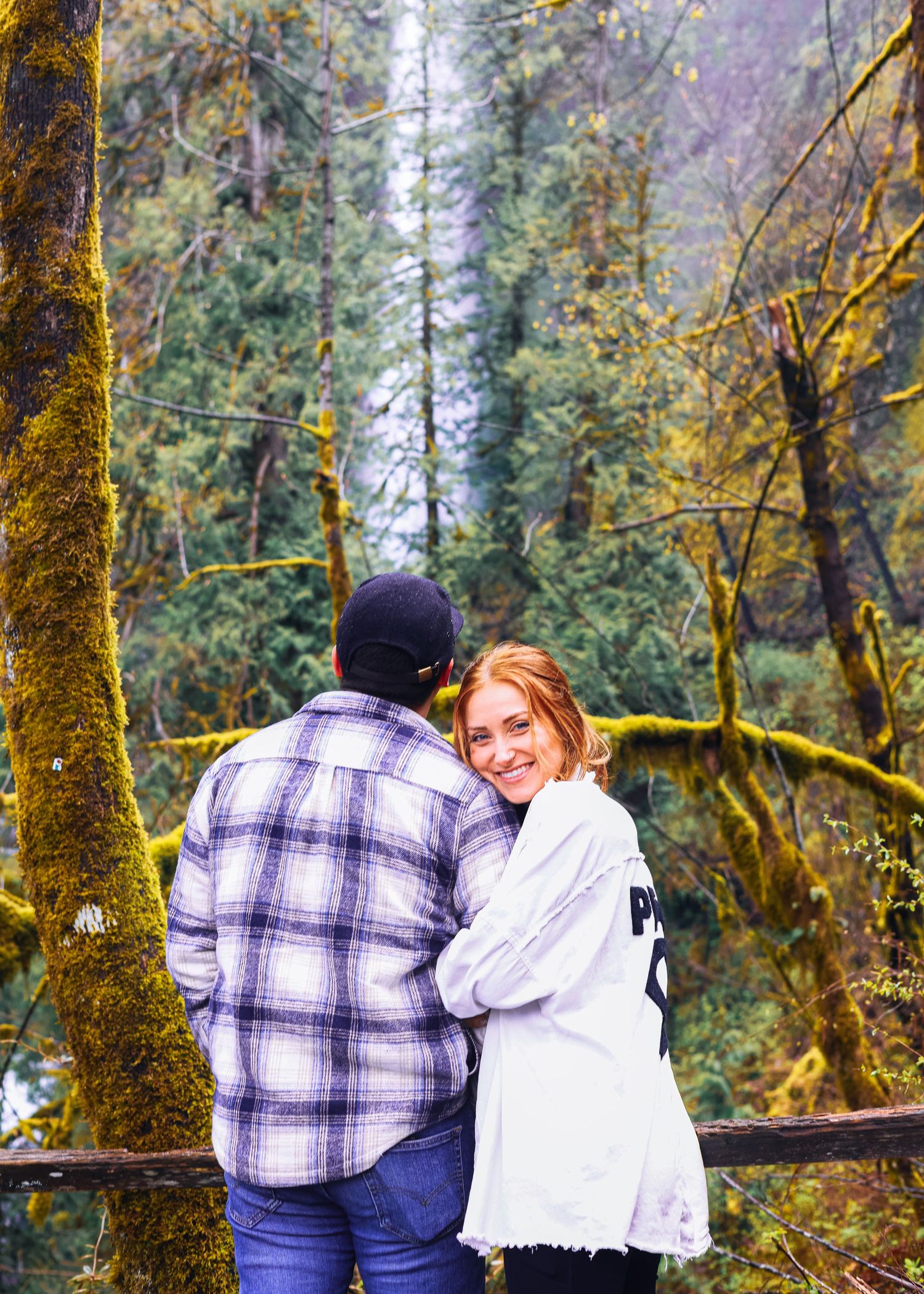 A smiling redhead woman leans on a man in a plaid shirt in a mossy forest.