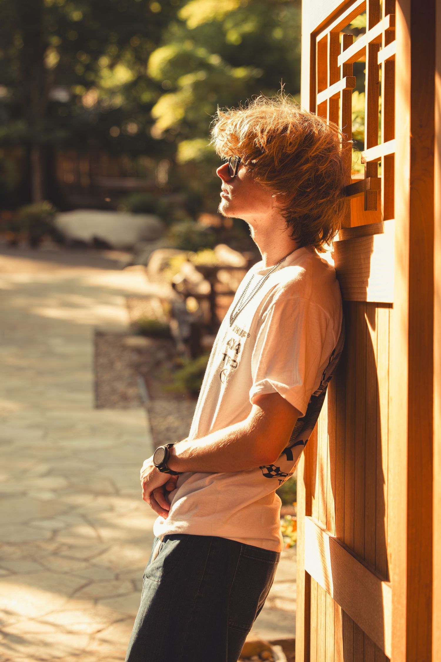 Curly redhead teen in white tee and sunglasses leans against wooden lattice wall in sunny garden.