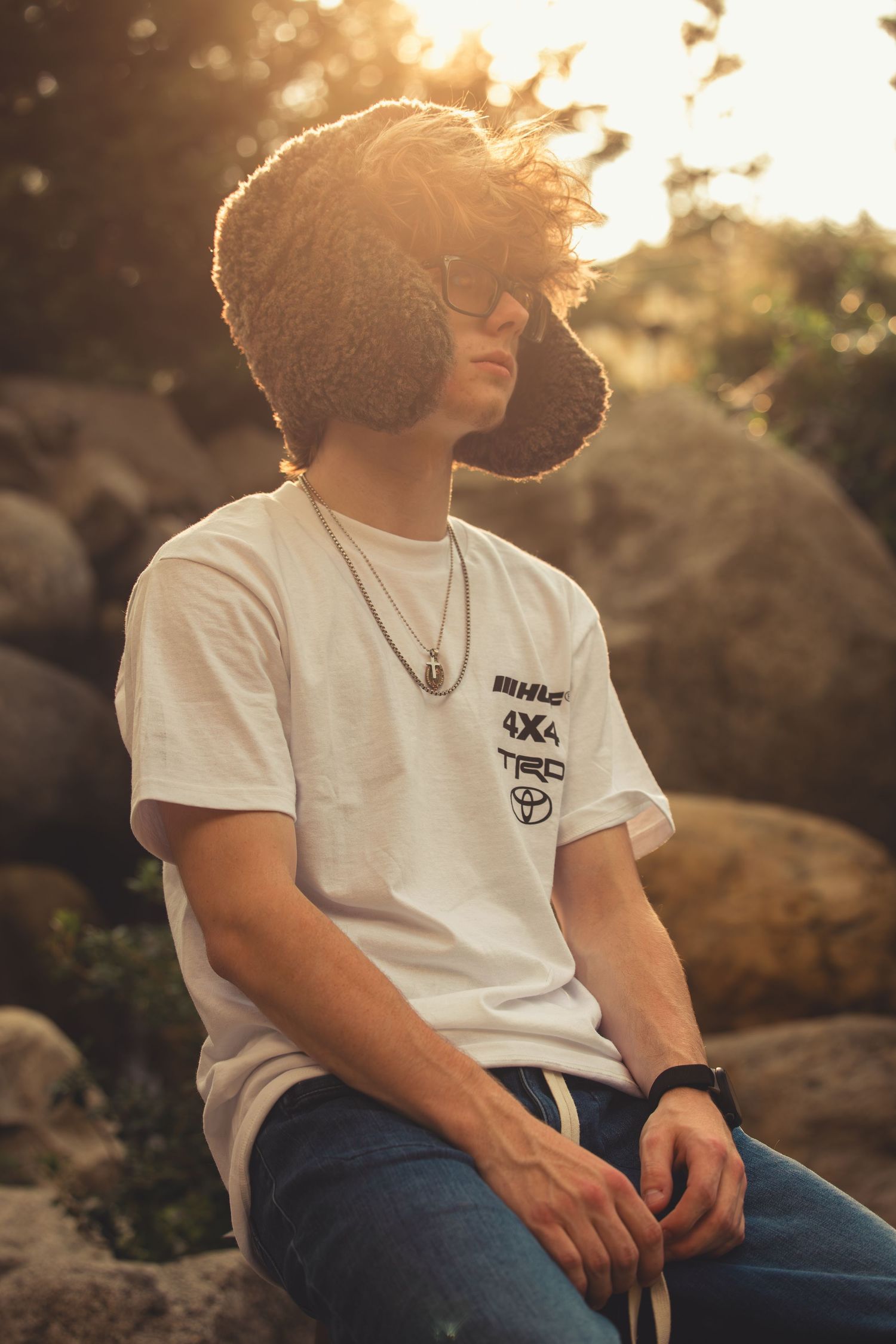 Young man in white Toyota TRD t-shirt wearing a fuzzy bucket hat sitting on rocks at golden hour.