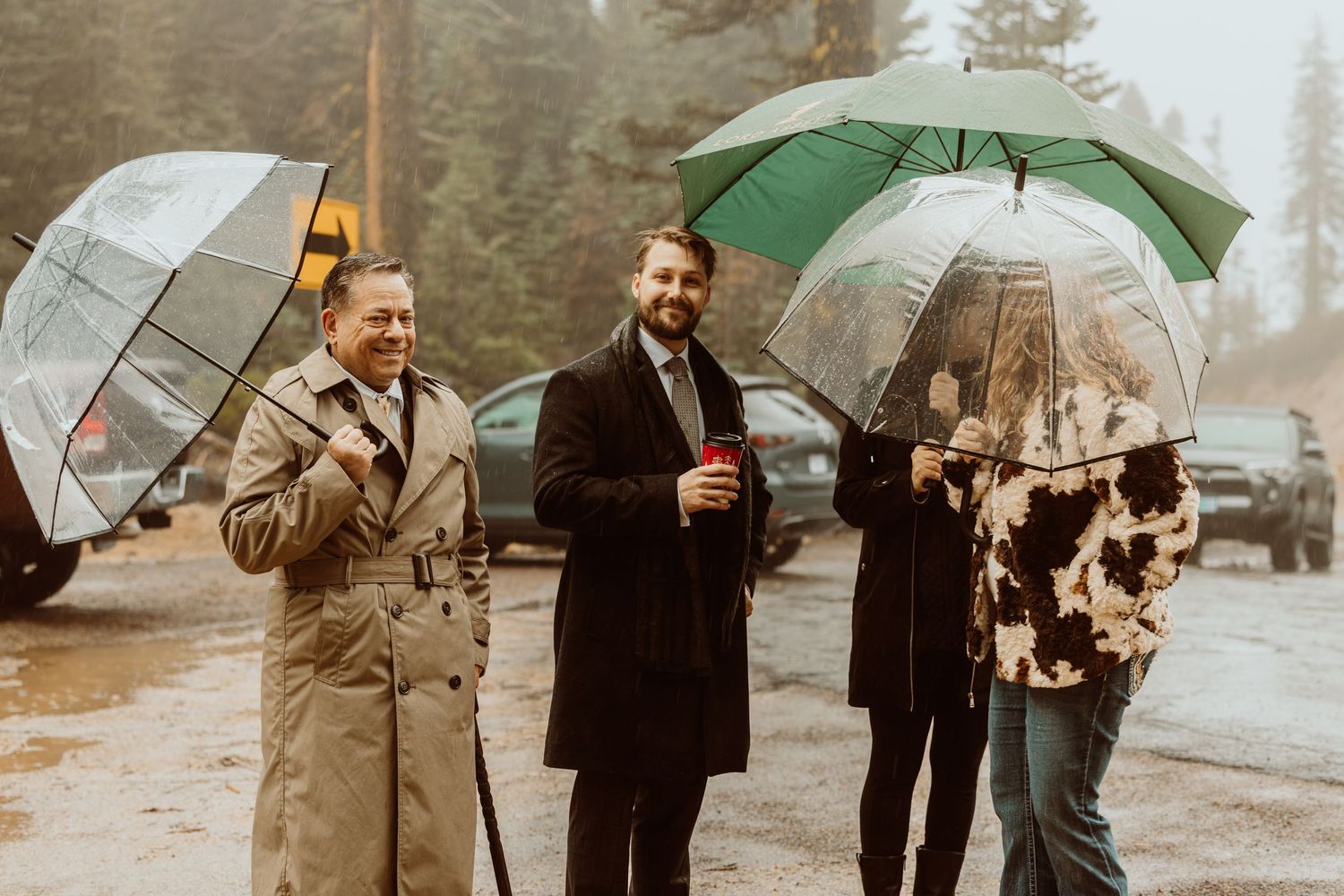 Two men in formal coats with umbrellas smiling outdoors on a rainy day.