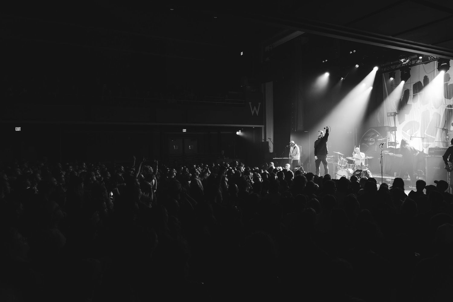 Black and white wide shot of large concert crowd facing illuminated stage during live music performance.
