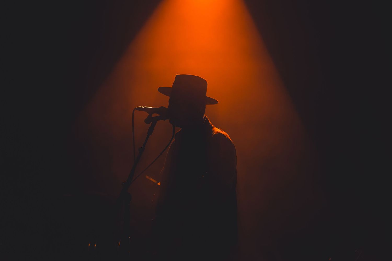 Silhouetted musician wearing a wide-brim hat performing at a microphone under dramatic orange stage lighting.