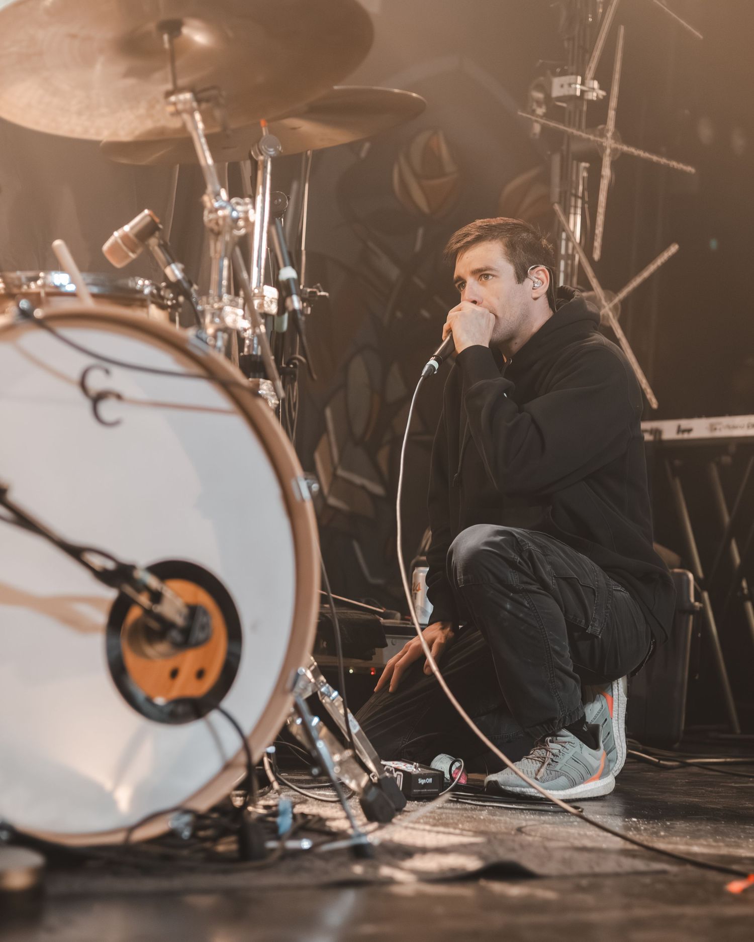 Male vocalist crouching on stage floor singing into microphone with drum kit visible in background during live concert.