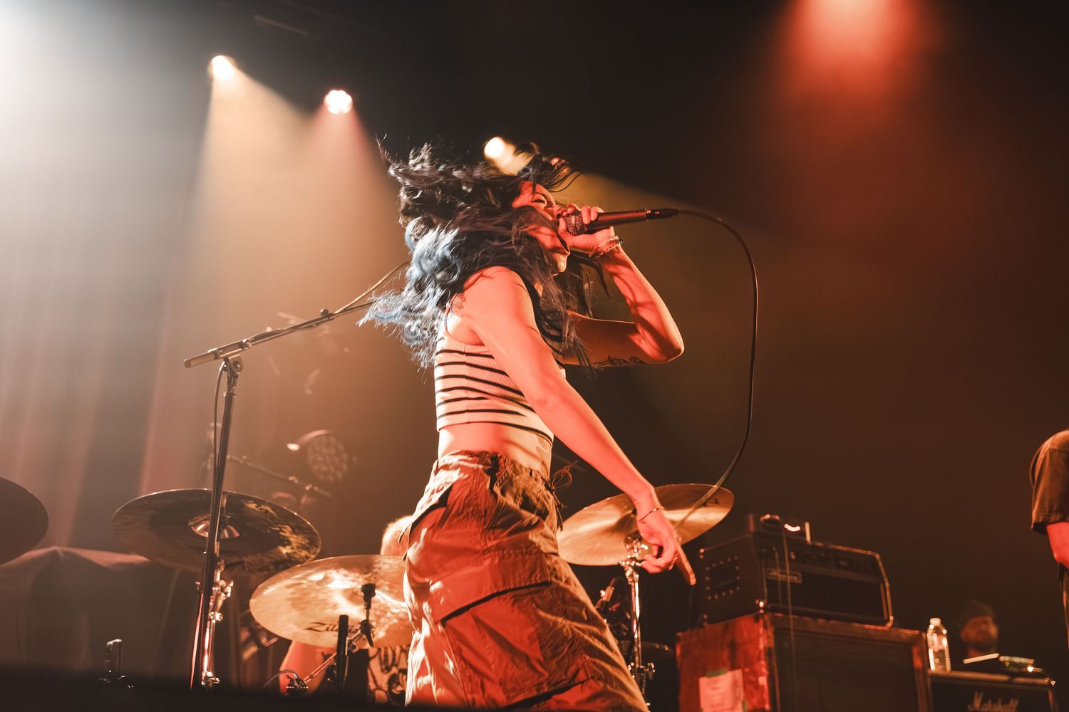 Female vocalist performing on stage under warm golden spotlights with drum kit and band visible in background.
