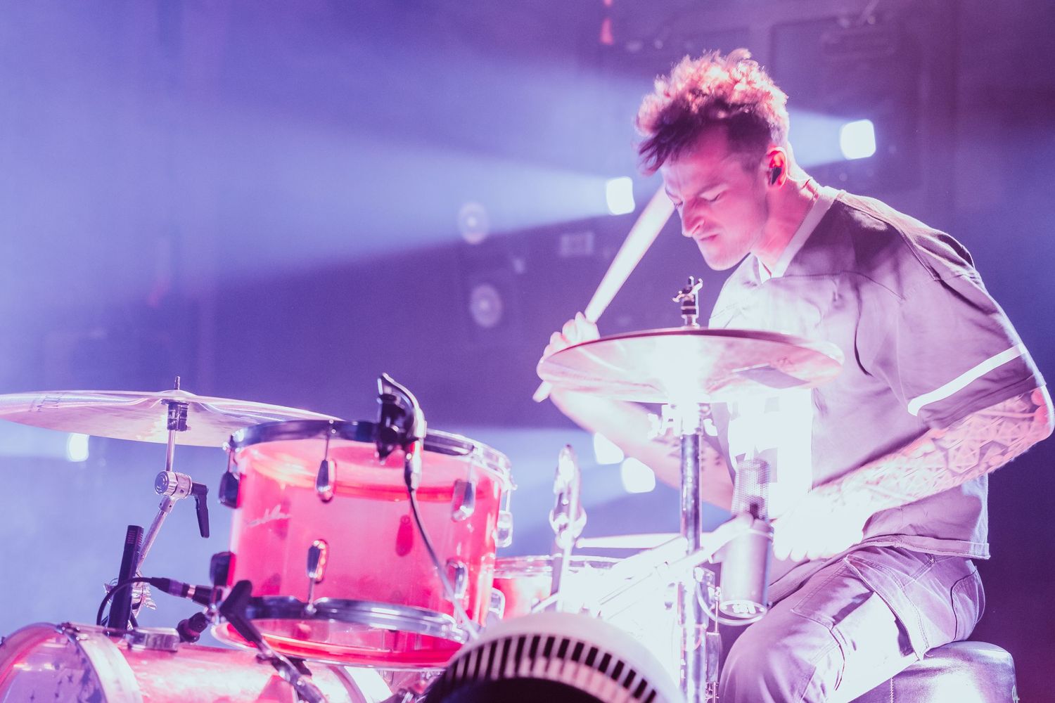 Male drummer with tattoos playing a red drum kit under pink and purple stage lights.