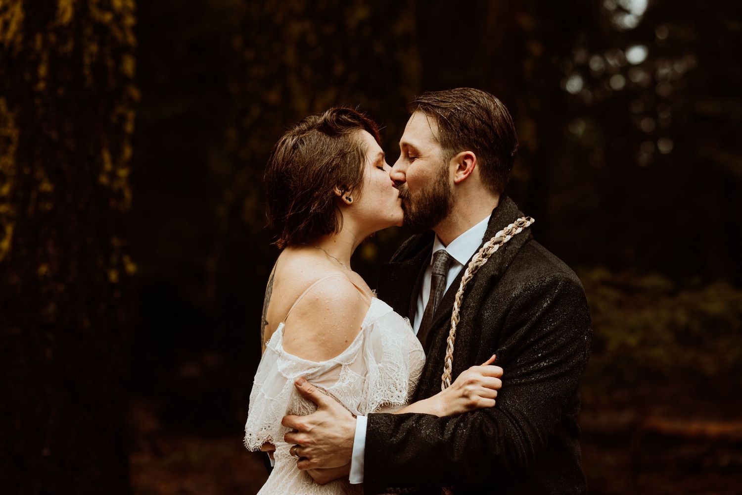 Bride and groom sharing a romantic kiss in a moody forest setting during their wedding day.