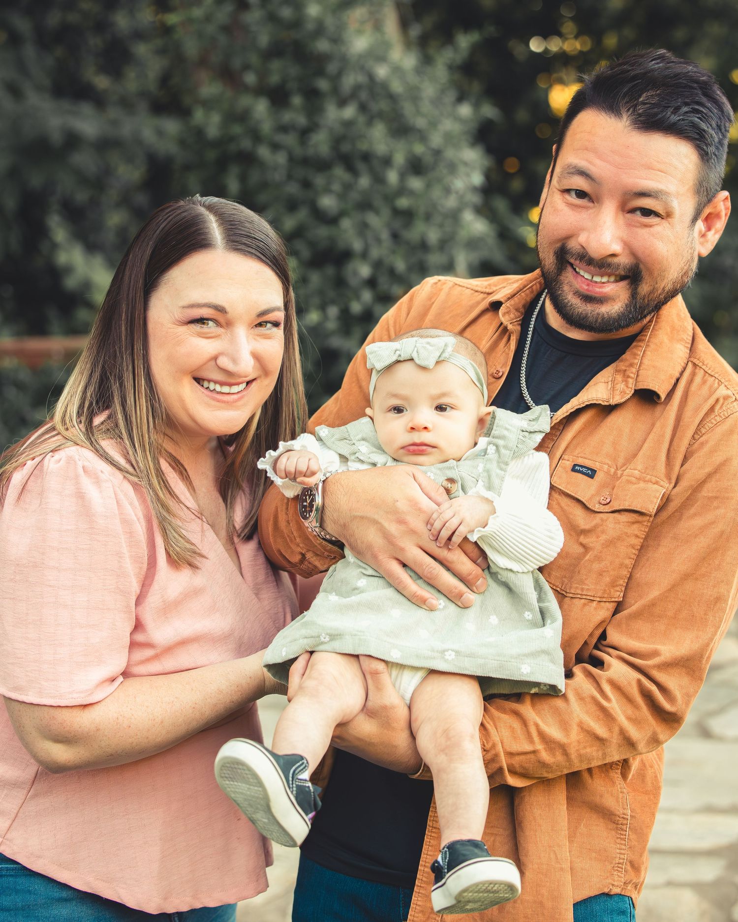 Happy parents posing with adorable baby girl in green dress outdoors among lush green bushes.