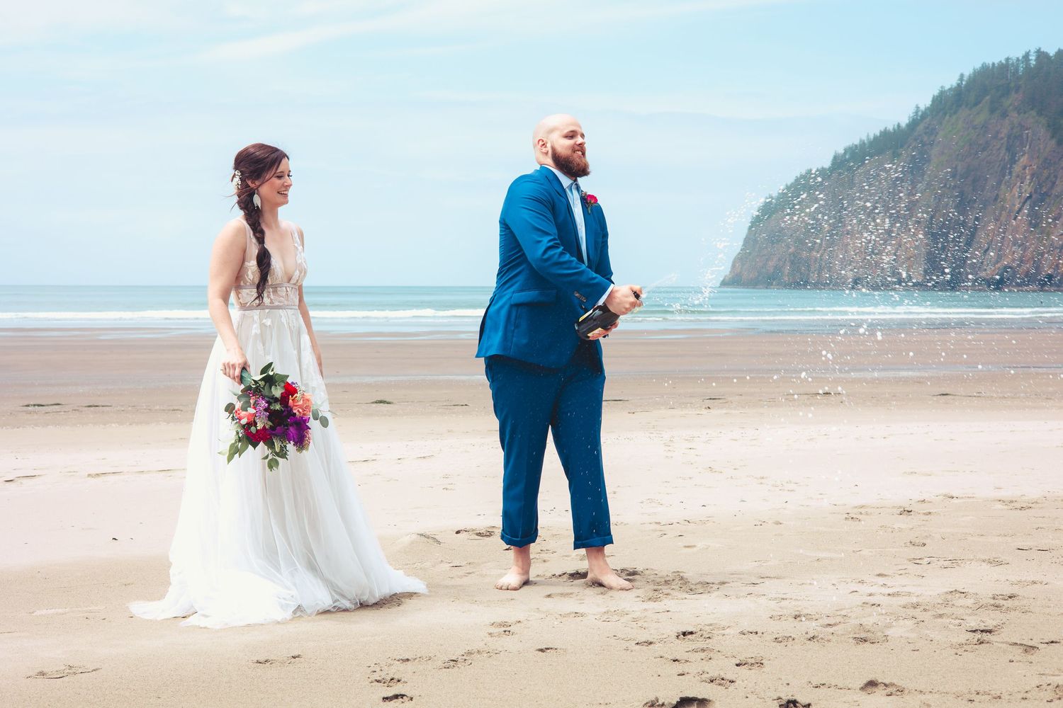 Bride and groom standing barefoot on a sandy beach with ocean waves and coastal cliffs behind them.