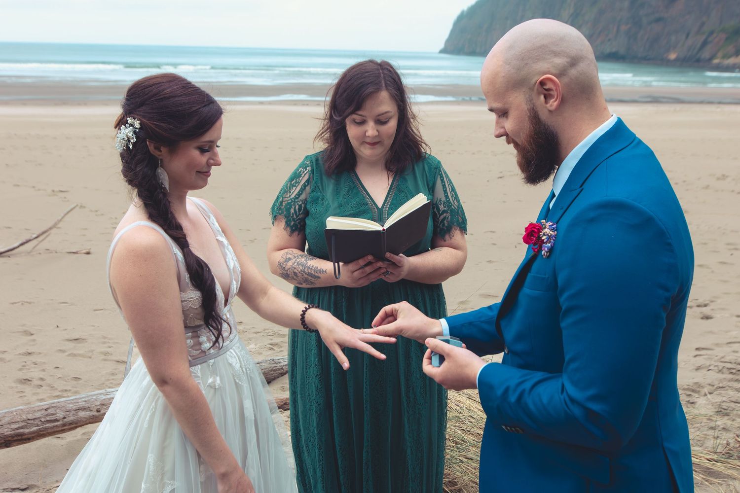 Groom places wedding ring on brides finger during an intimate beach ceremony with officiant present.