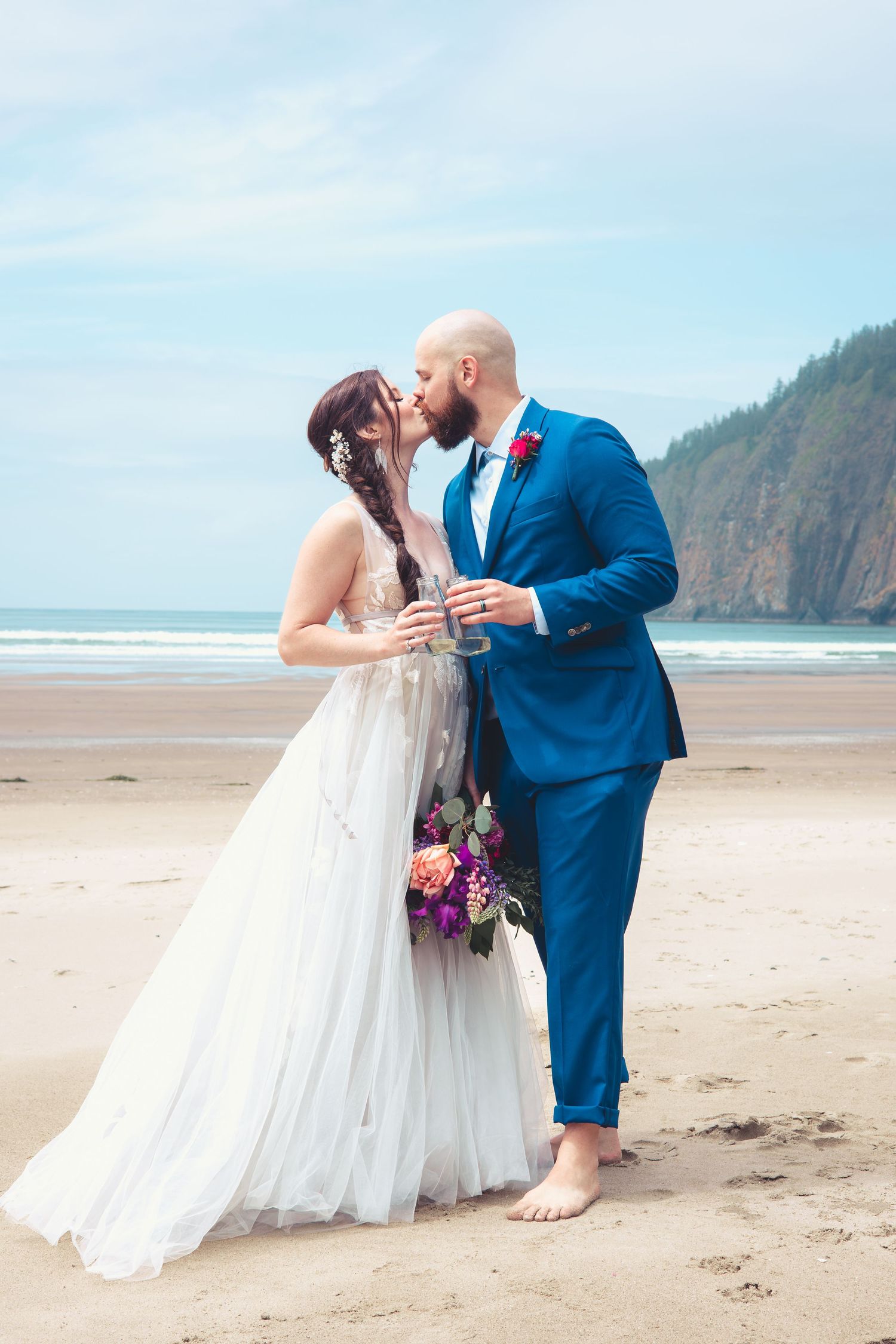 Bride and groom sharing a kiss while holding champagne glasses on a scenic coastal beach.