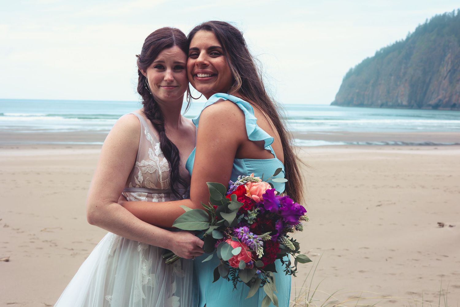 A bride in a white gown and her bridesmaid in a teal dress smile together on a scenic beach.