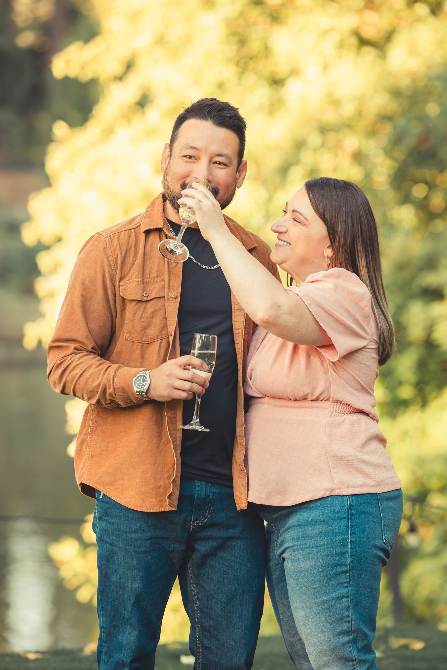 Laughing woman playfully feeding champagne to her partner outdoors in a golden autumn park.