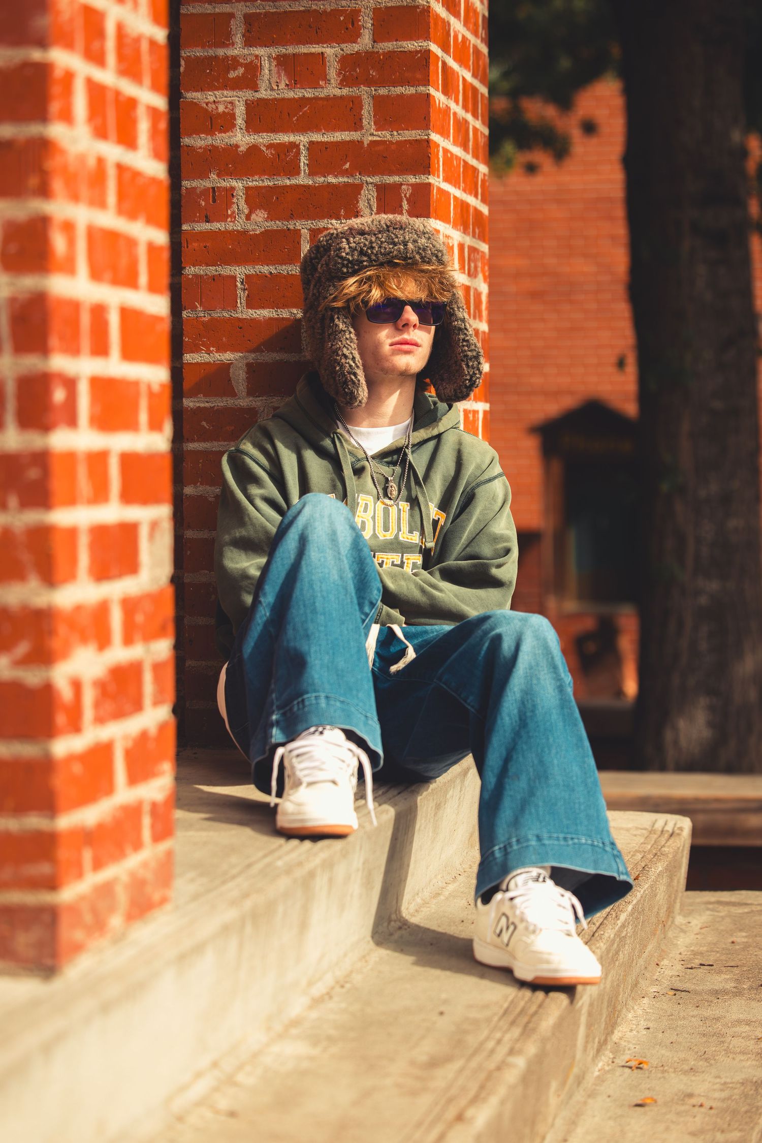 Teen in Humboldt State hoodie and fur trapper hat sits on concrete ledge against red brick wall.
