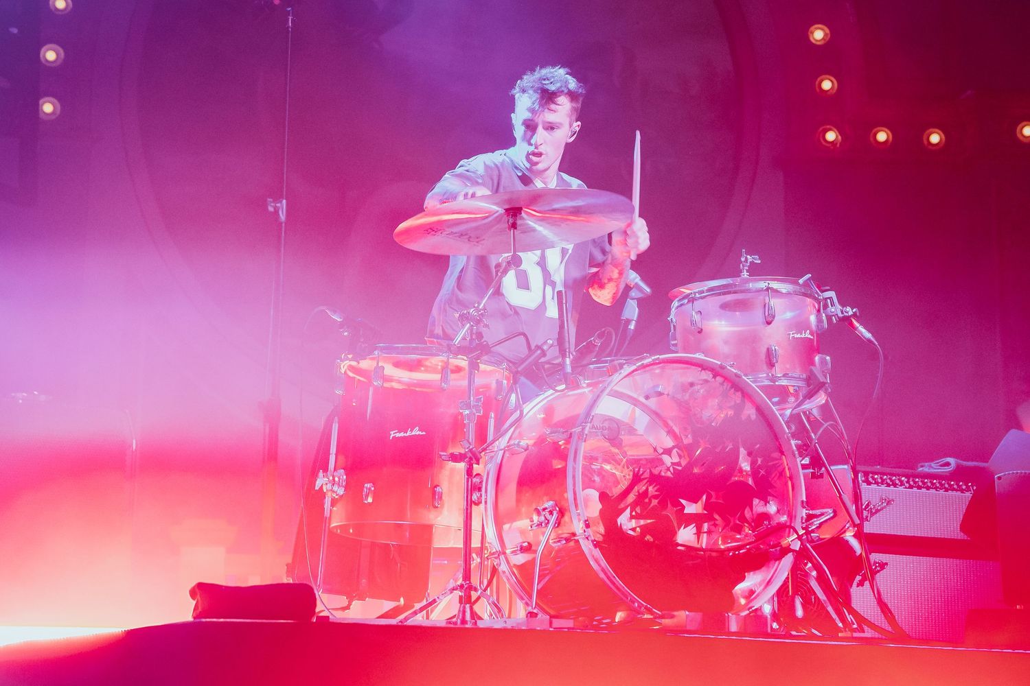Drummer performing energetically on stage surrounded by warm pink and red concert lighting.