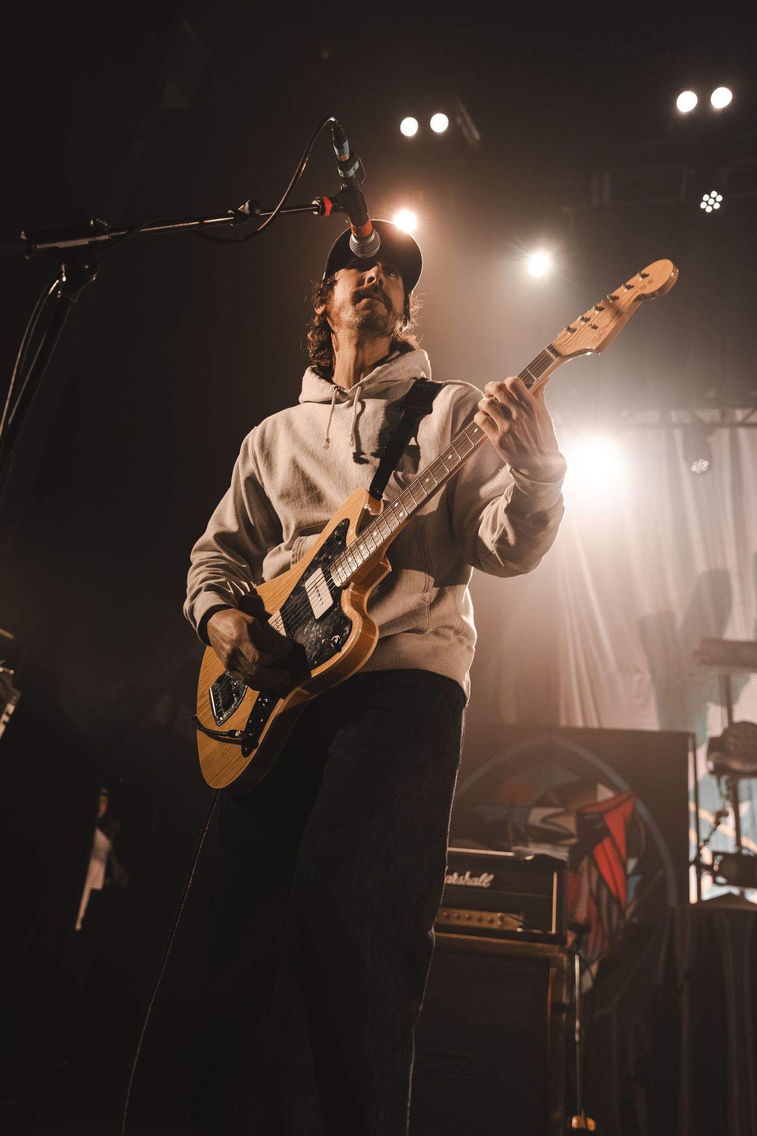 Guitarist in white hoodie and cap playing electric guitar on stage under warm concert lighting.