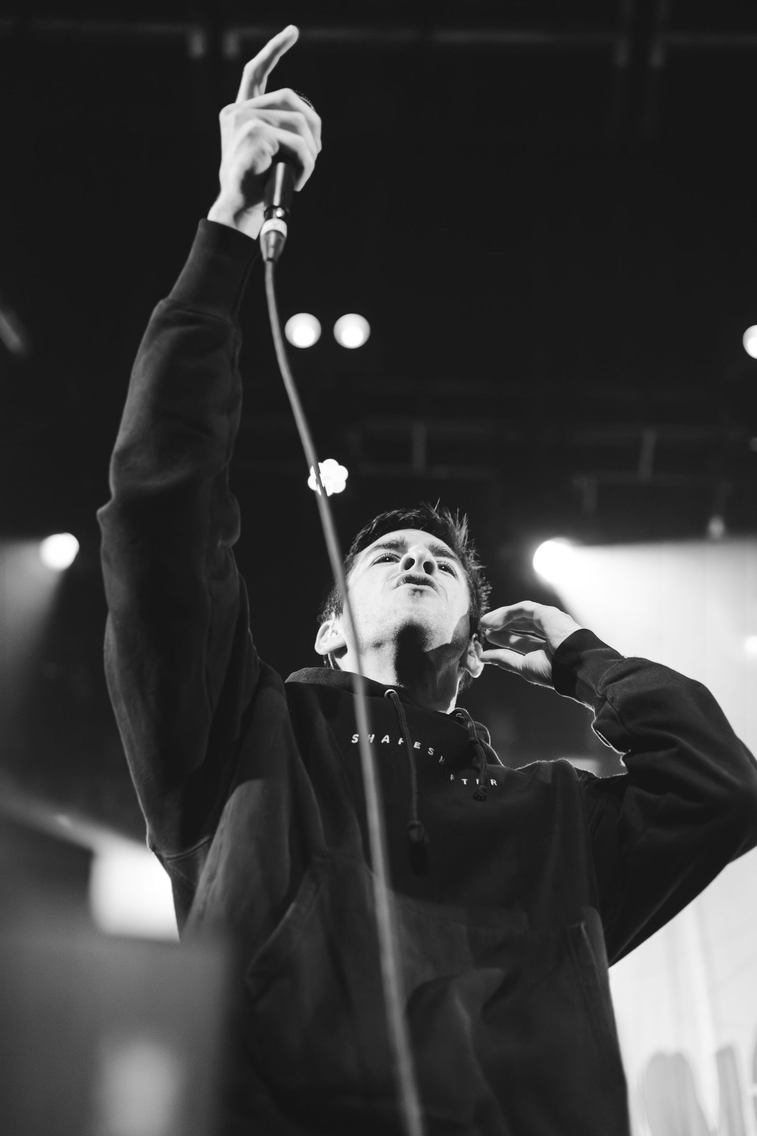 Black and white low-angle shot of a vocalist pointing upward dramatically during a live performance.