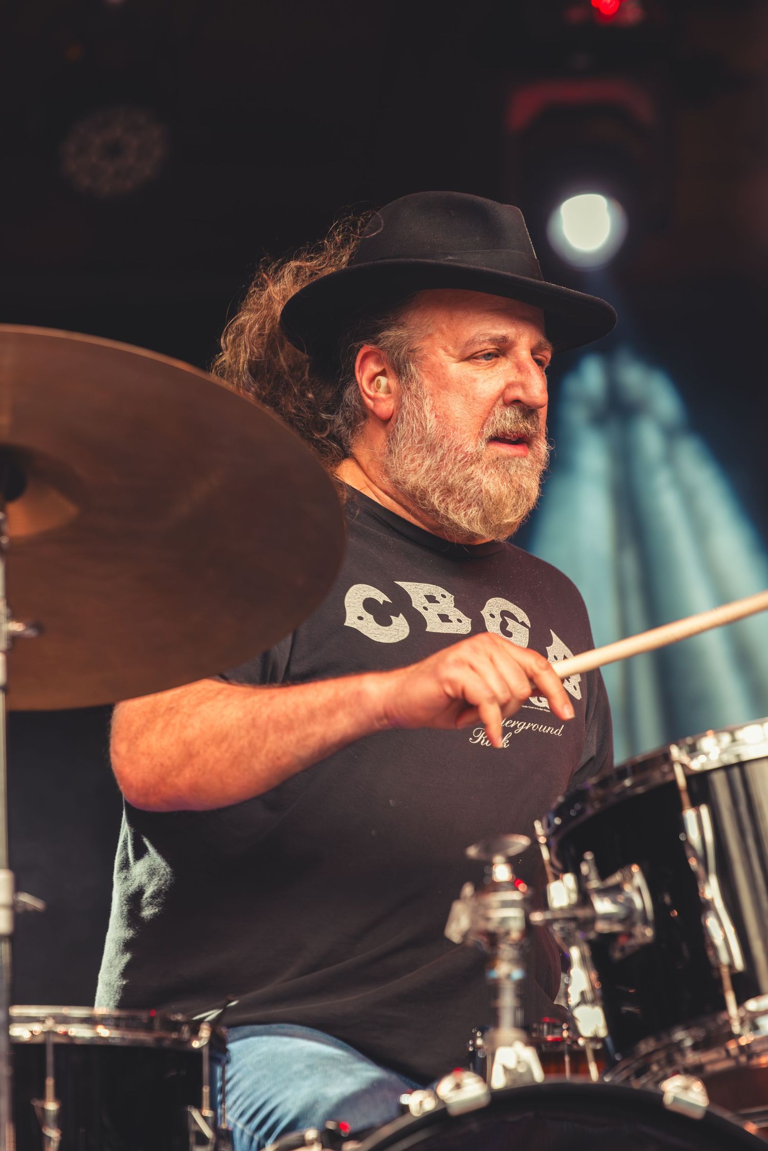 Bearded drummer in a black hat playing drums at a live music concert.