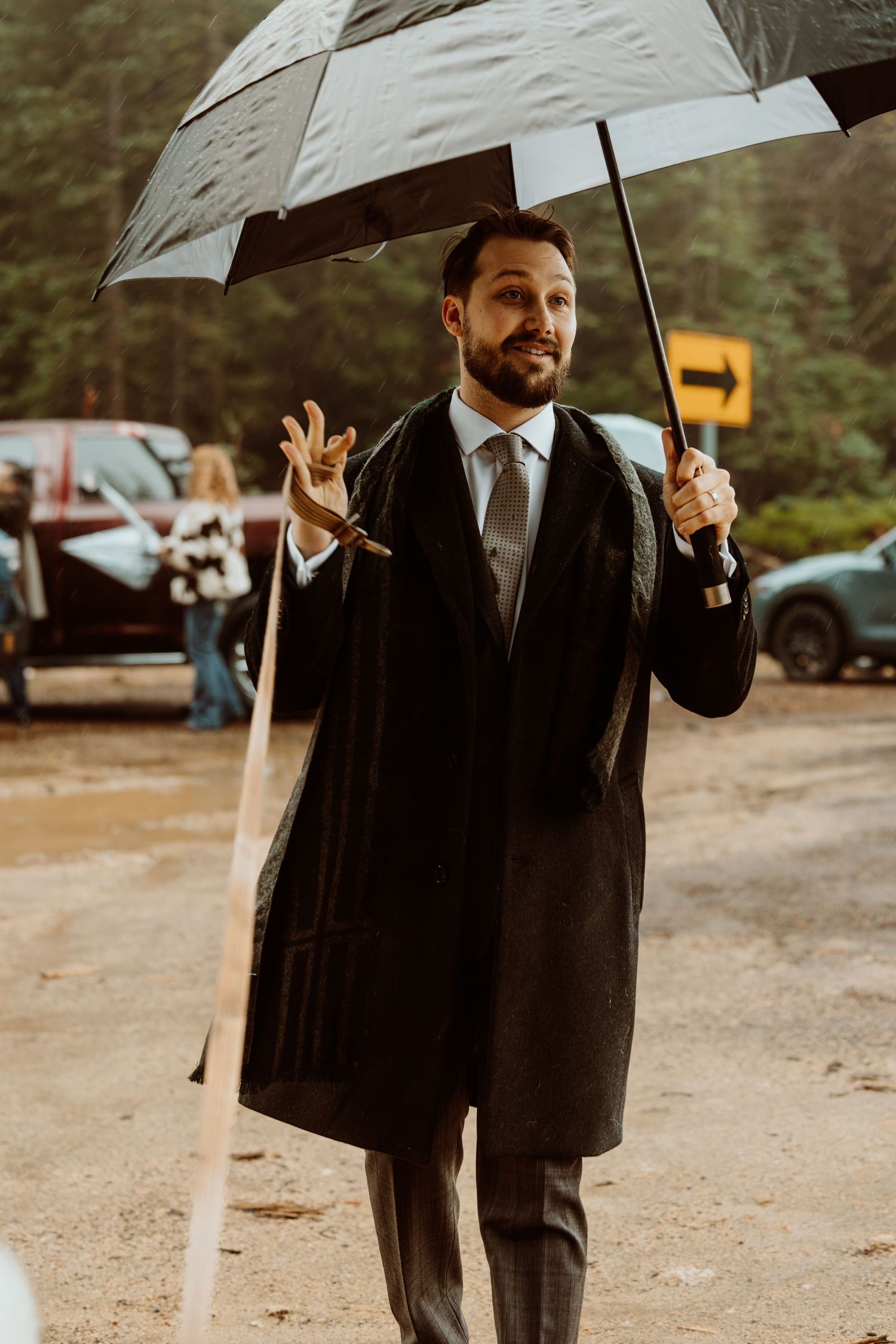 Dapper bearded man in long dark coat holds umbrella while gesturing outdoors in rainy forest setting.