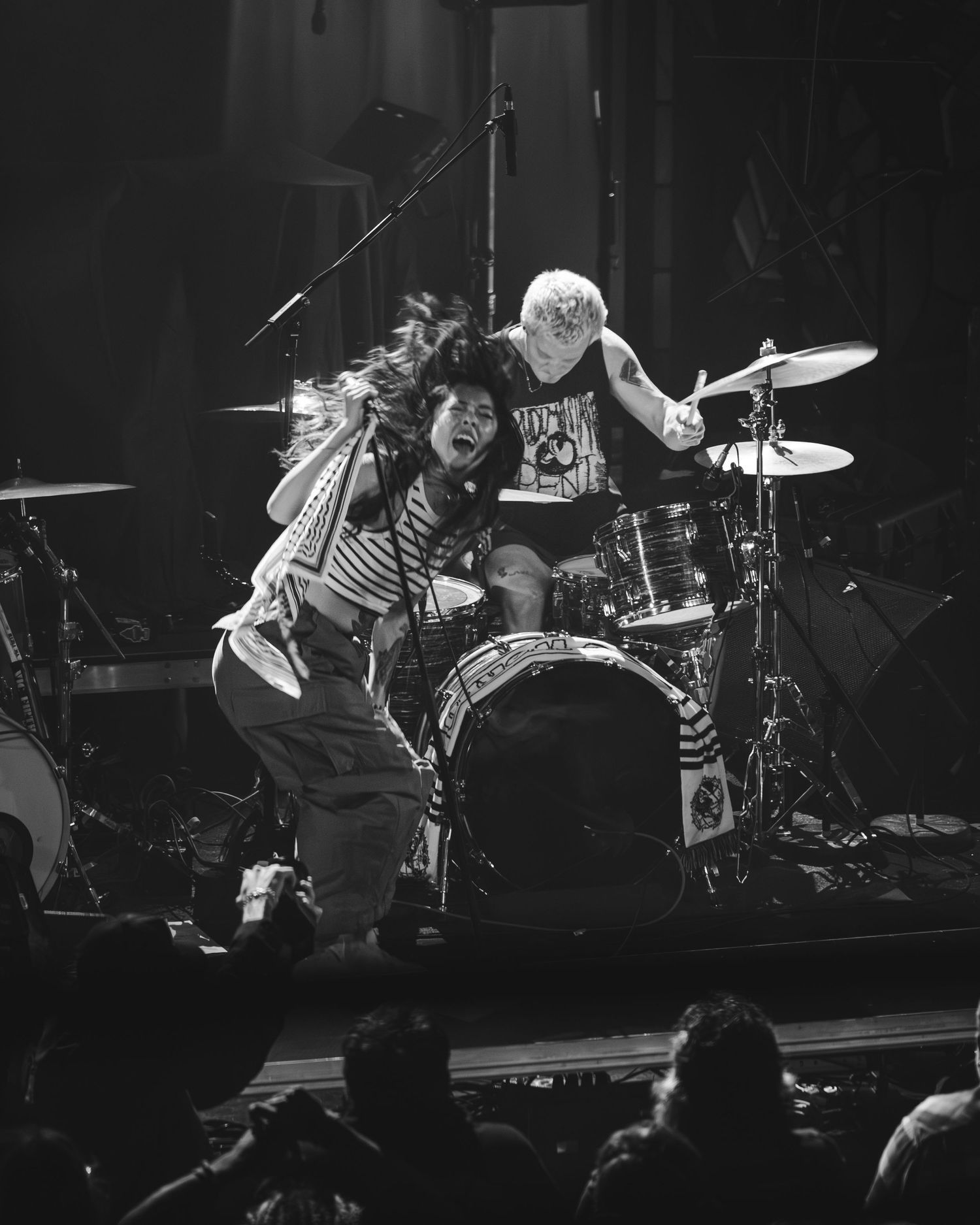 Energetic black and white concert photo of a vocalist leaning toward crowd with drummer performing behind on stage.