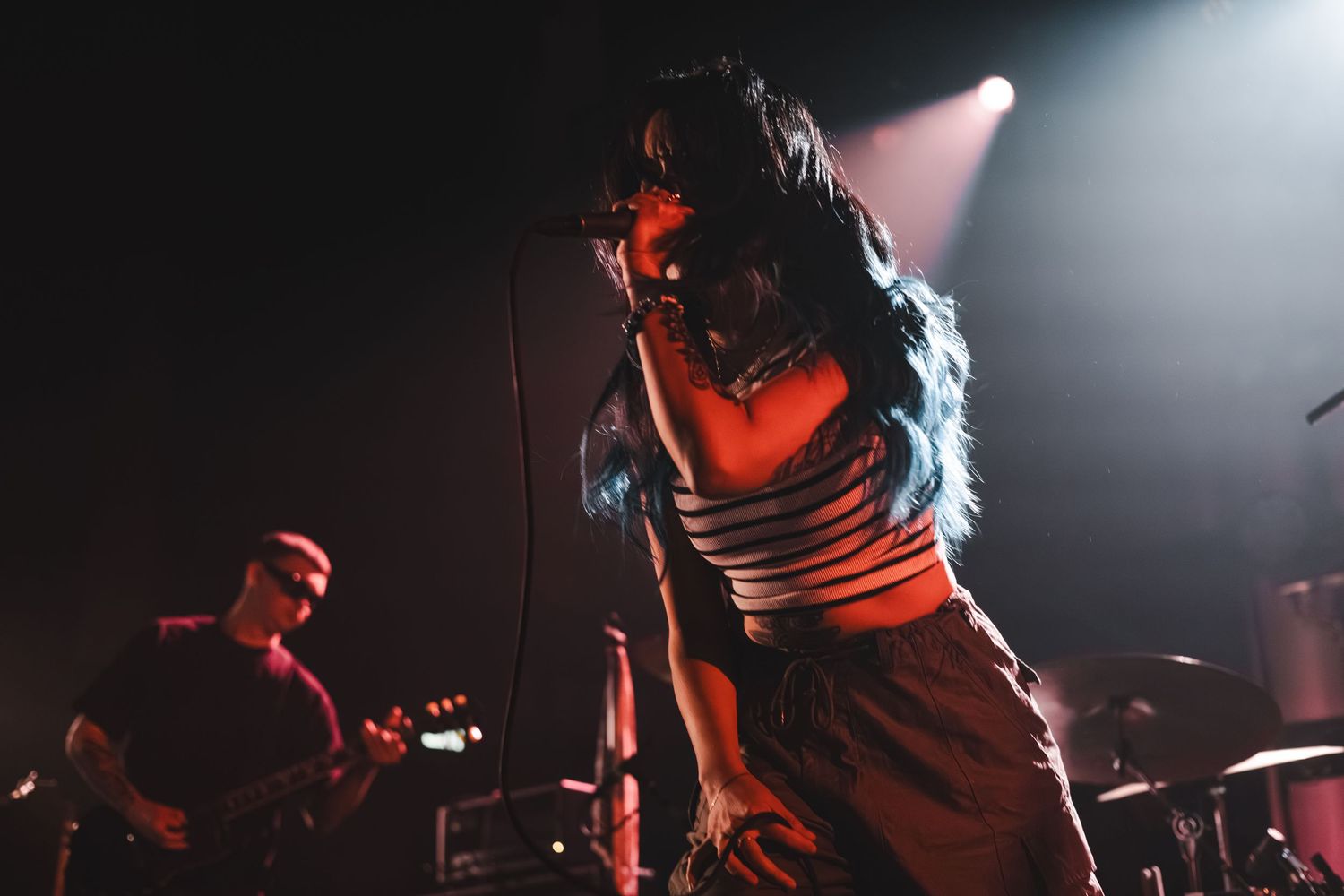 Female vocalist leans forward singing passionately on stage with guitarist visible in background.