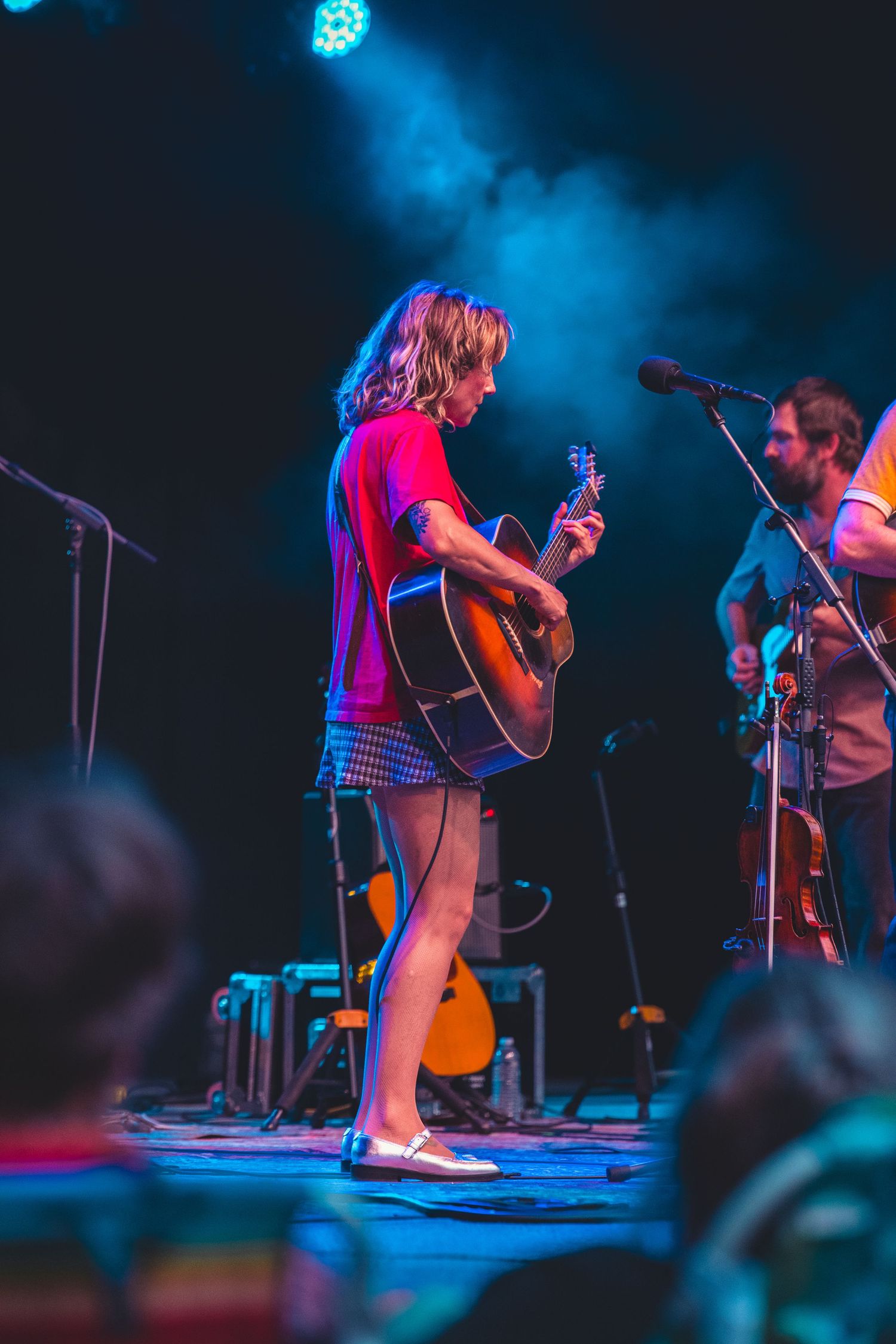 Woman in red shirt playing acoustic guitar on a colorful blue and red lit concert stage.