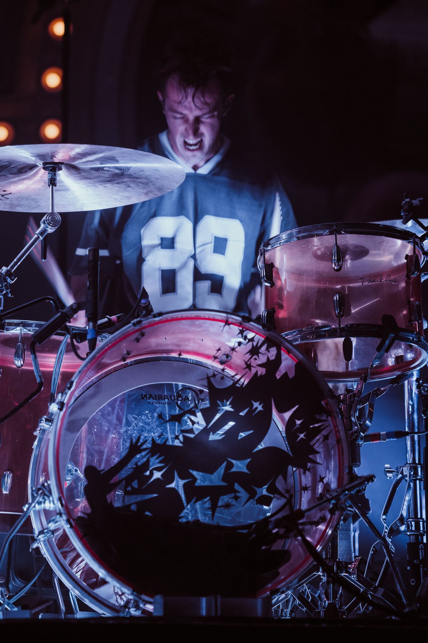 Energetic drummer plays drum kit on stage wearing number 89 jersey at live concert.