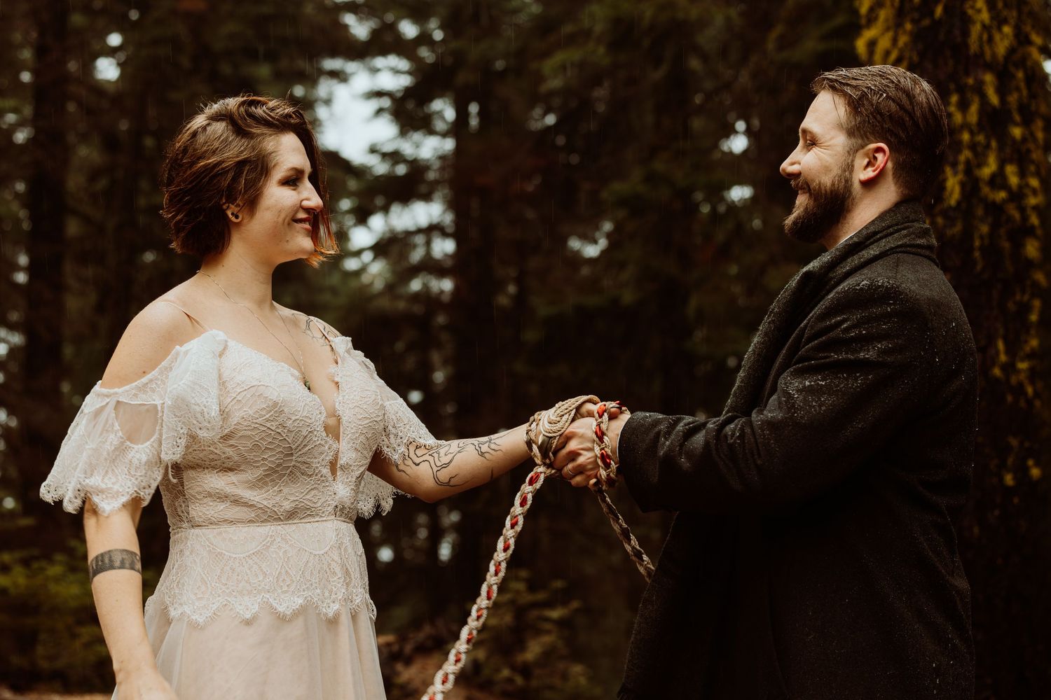 Smiling bride in lace dress and tattooed groom holding hands during an outdoor forest wedding ceremony.