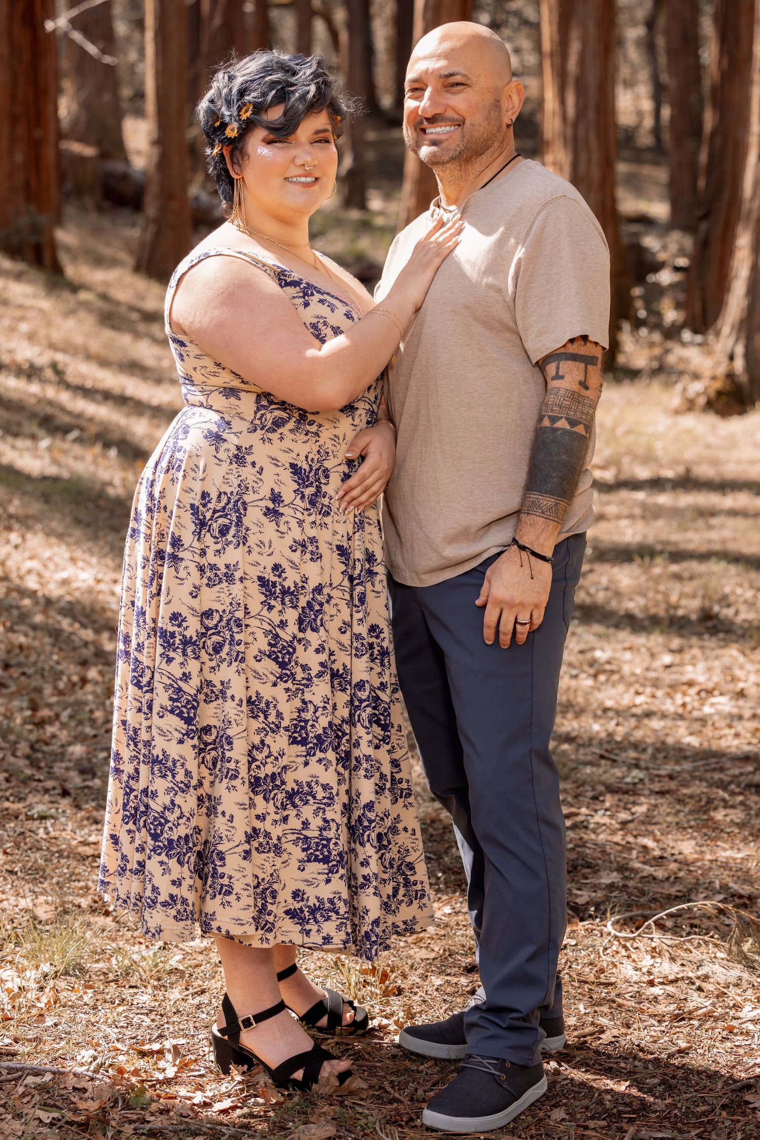 Couple posing in forest, woman in floral dress with man in beige shirt and navy pants.
