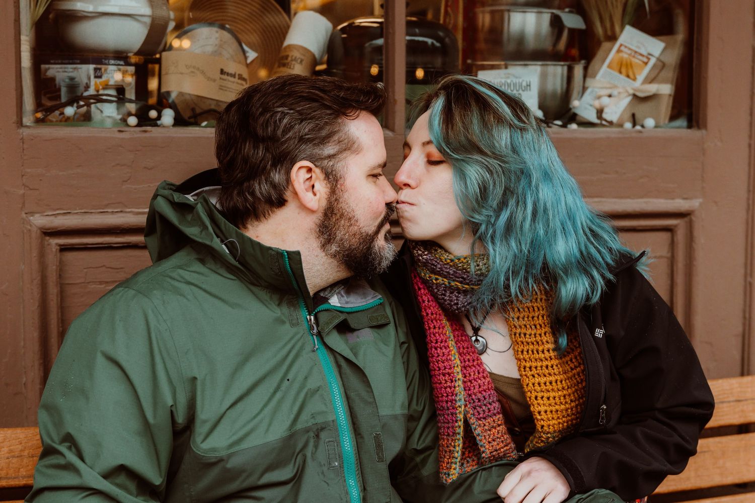 Couple sharing a romantic kiss on a bench outside a vintage storefront.
