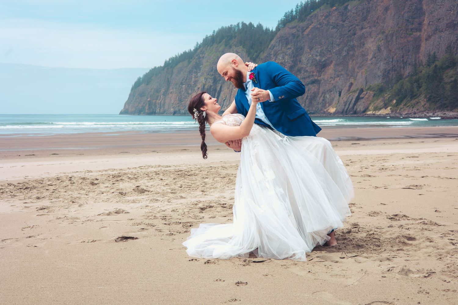 Groom dips and kisses bride on sandy beach, white gown flowing, rocky tree-covered cliffs in background.