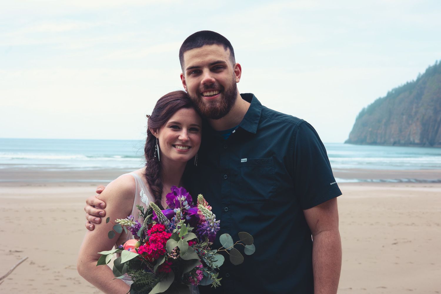 A smiling bride holding a purple and pink bouquet poses with a man in a dark shirt on a beach.