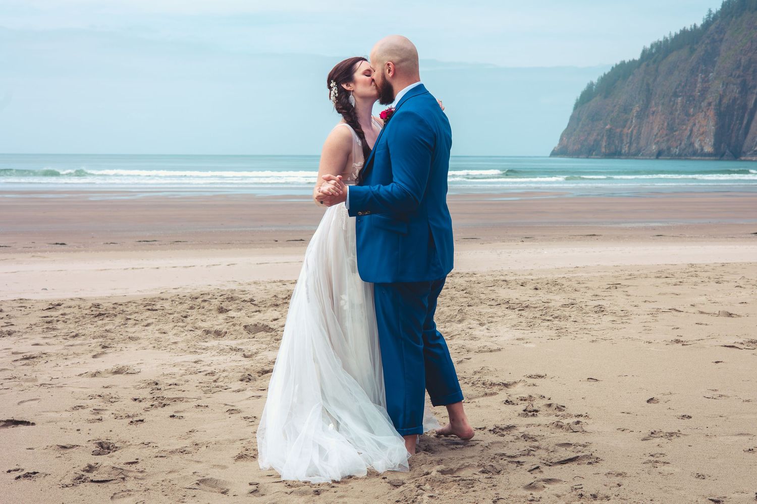 Bride and groom share a romantic kiss on sandy beach, groom in blue suit, rocky cliffs in background.