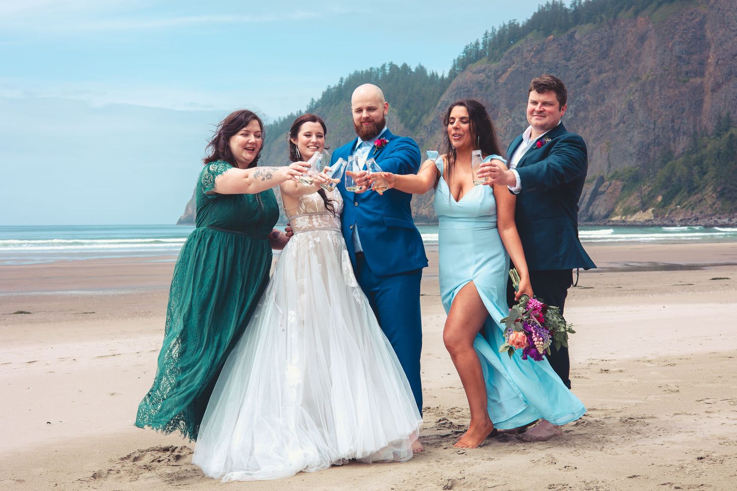Wedding party of five toasting with drinks on a sandy beach with green forested cliffs behind.