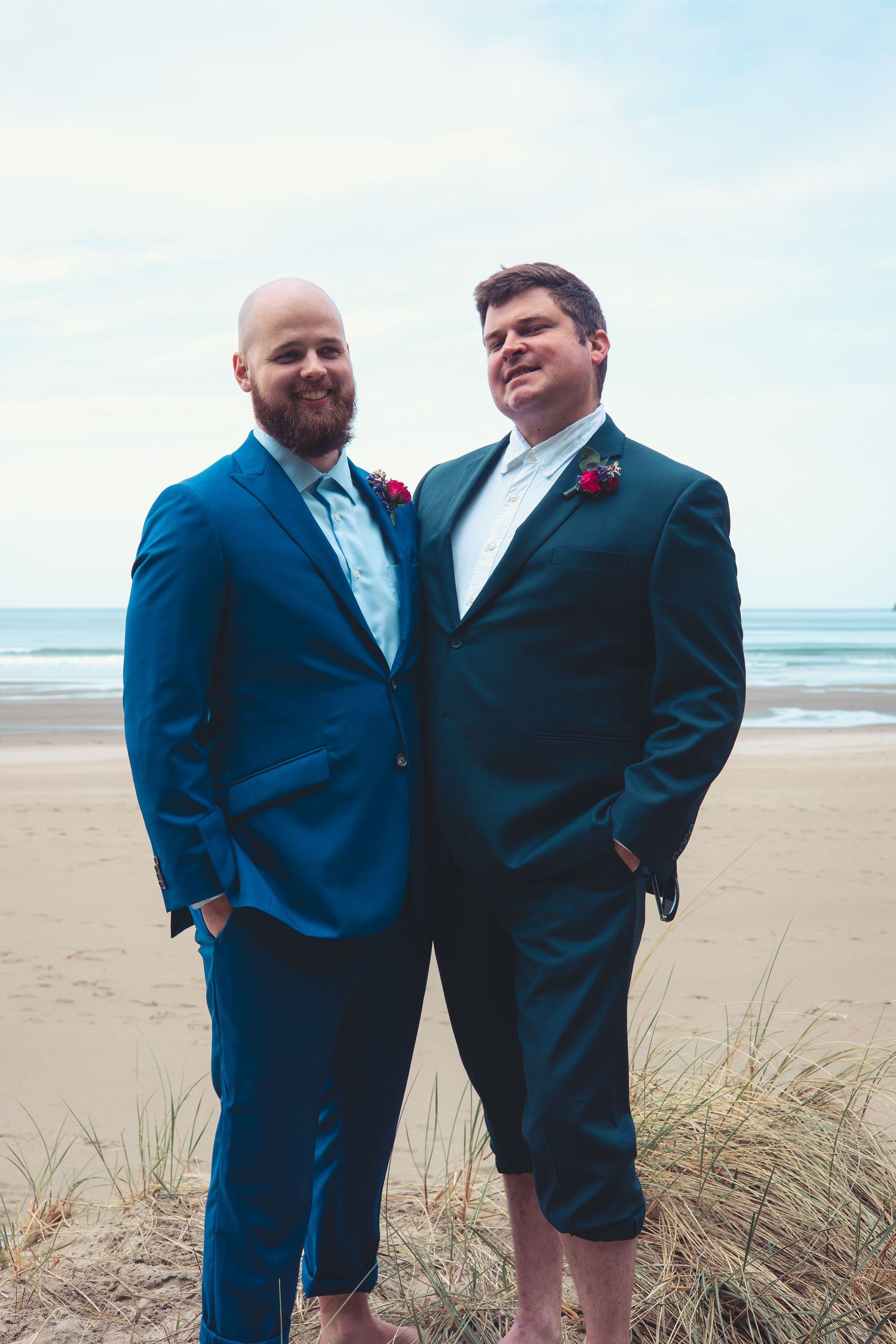 Two grooms in blue suits pose together on a sandy beach on their wedding day.