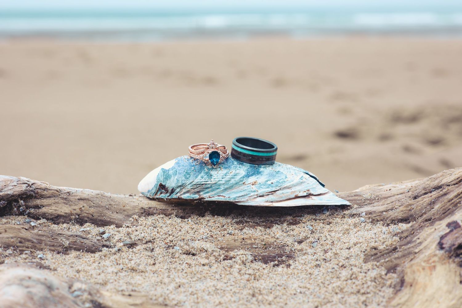 Wedding rings displayed on blue fabric resting on driftwood at a serene sandy beach.