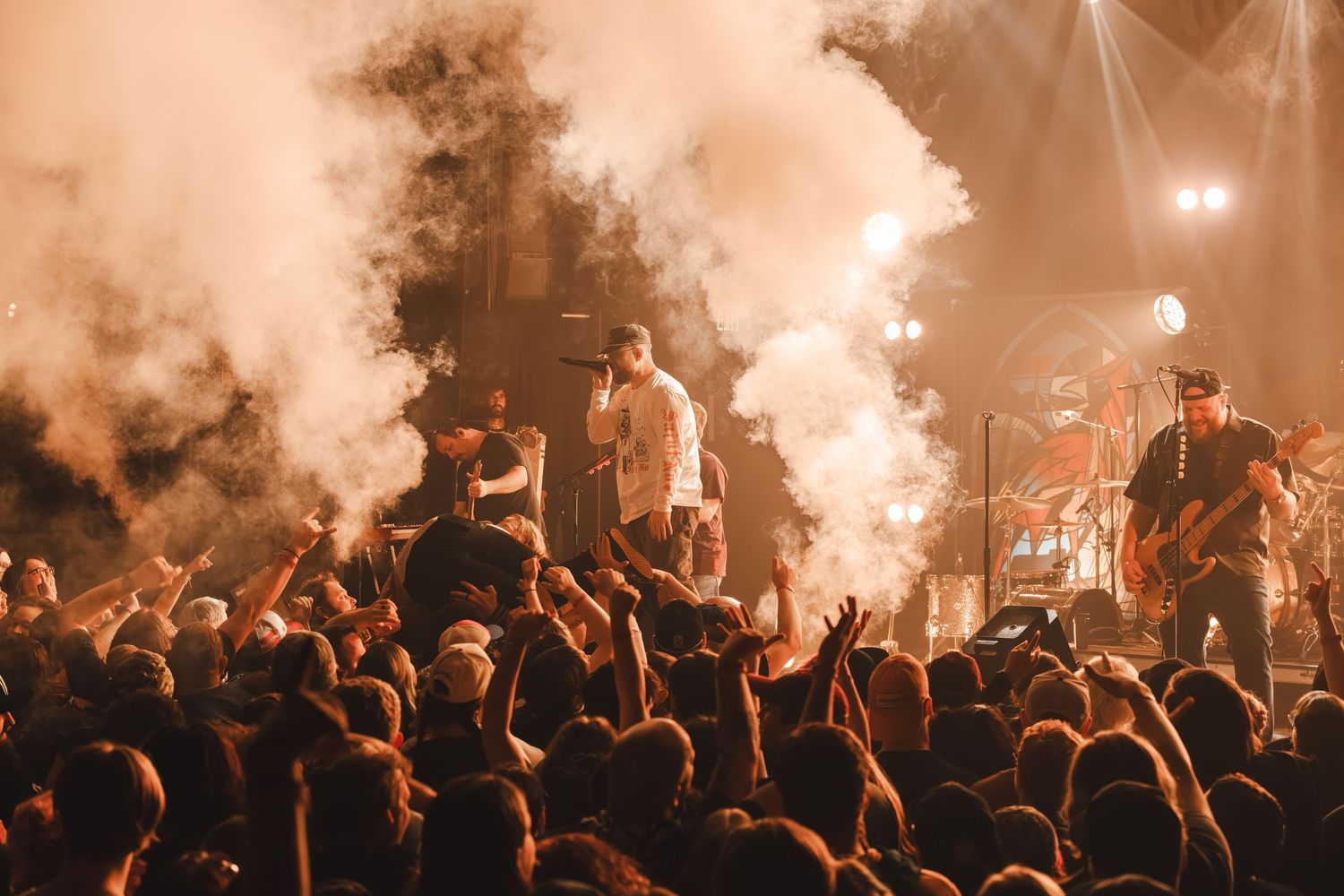 Band performing on stage surrounded by dramatic smoke and warm orange lighting with crowd.