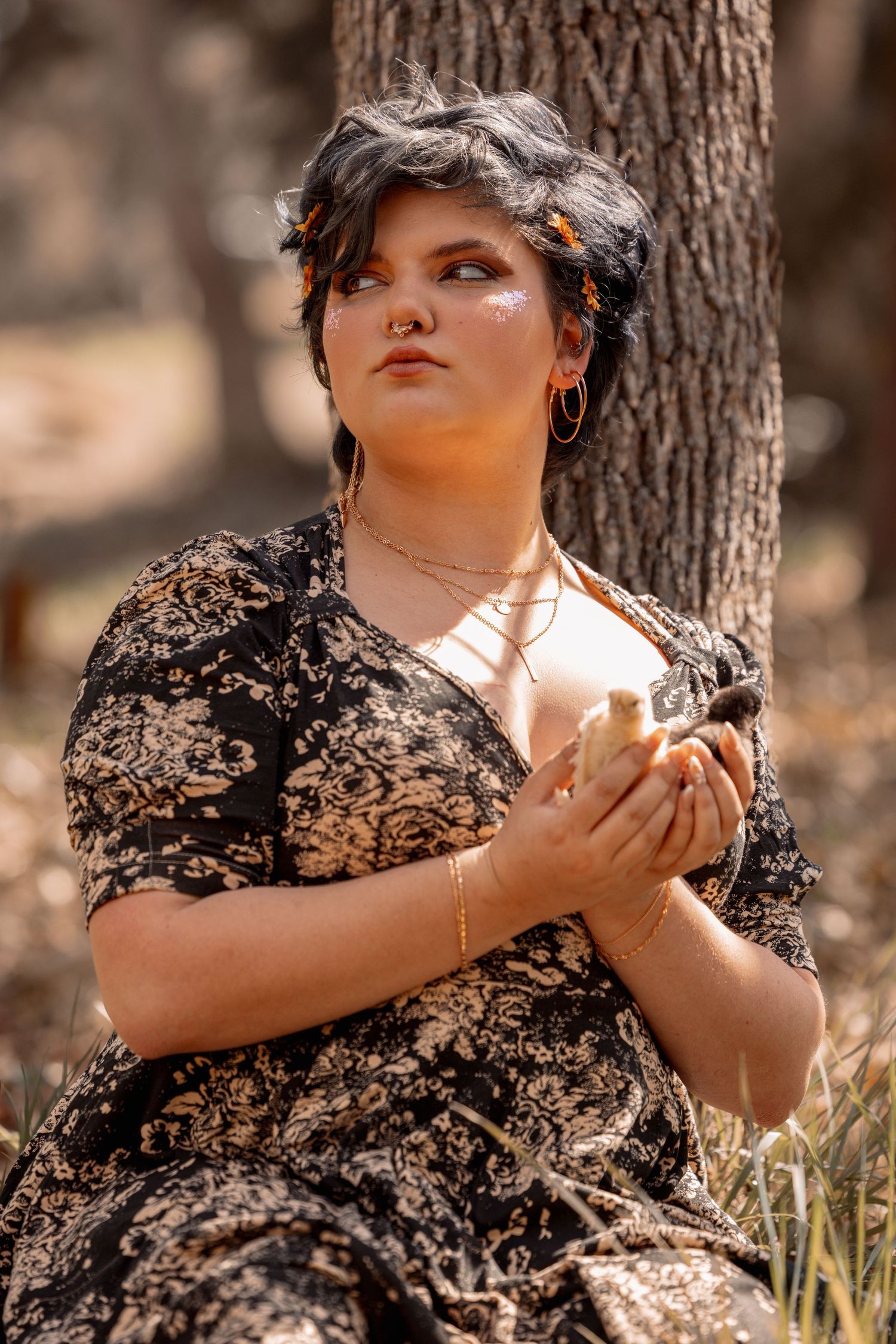 Woman in floral dress holds a baby chick close to her chest while sitting against a tree outdoors.