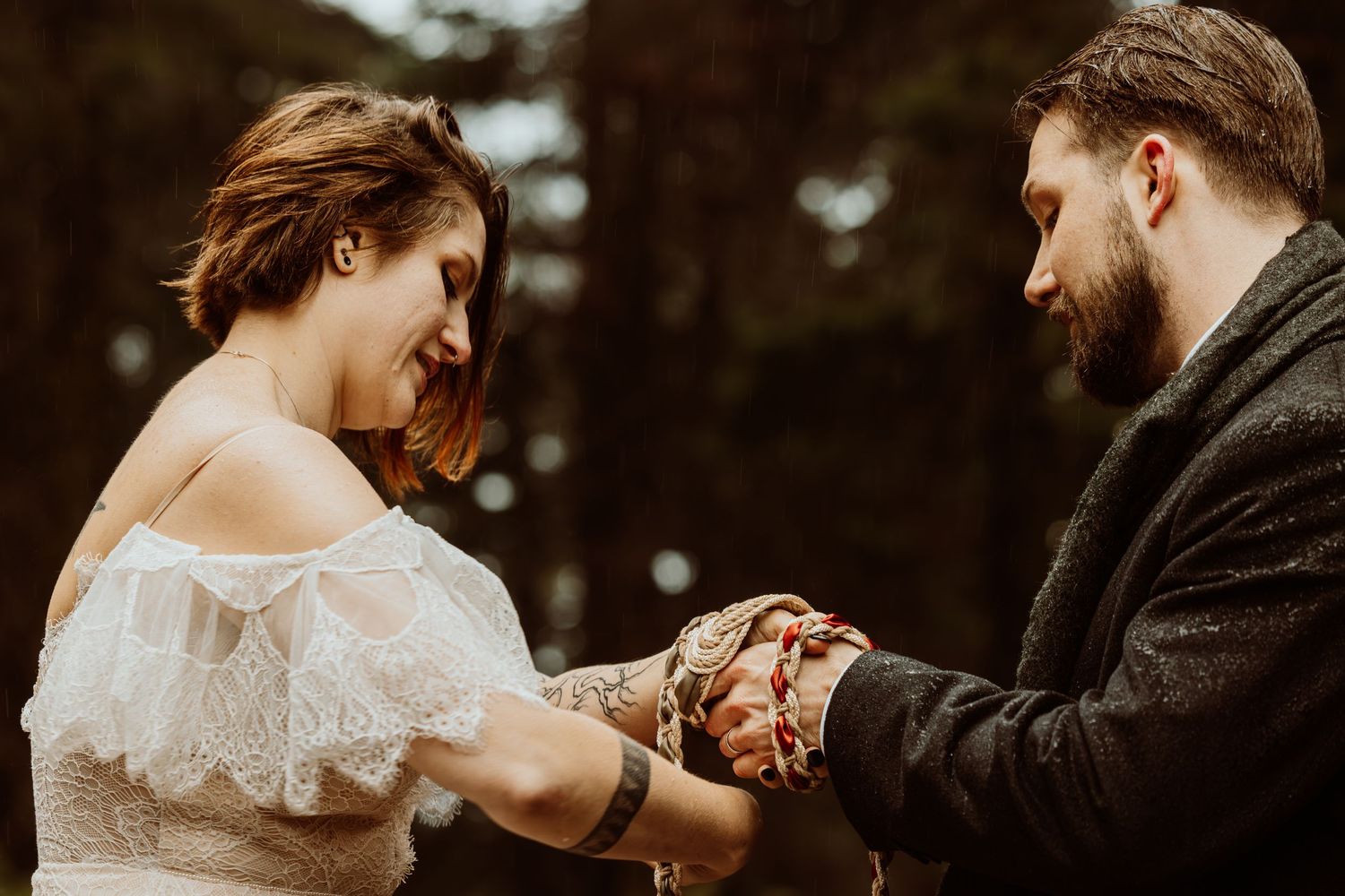 Bride and groom share tender handfasting ritual with red braided cord during outdoor forest wedding.
